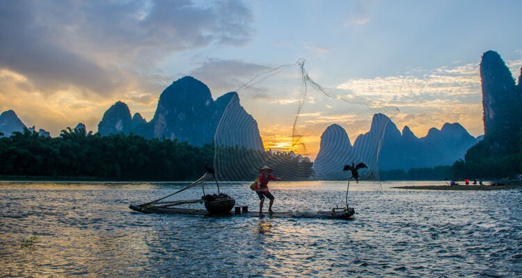 An old man on a bamboo raft is casting a net to catch fish on the Li River at sunset