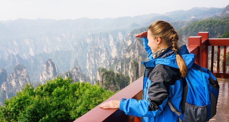 A young woman wearing a blue windbreaker and carrying a backpack happily leaned against the railing, gazing at the scenery of Zhangjiajie National Forest Park on a sunny day, shielding her eyes from the sun with one hand.