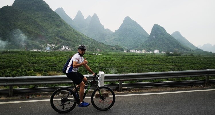 An international traveler is riding a bicycle on a rural road in Yangshuo and giving a thumbs-up to the camera.